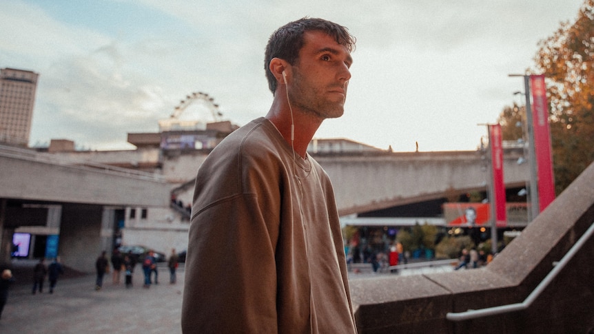 A man stands in a concrete cityscape with white headphones in and a grey shirt on.