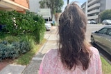 The back of a brown-haired woman standing on the footpath along a street.