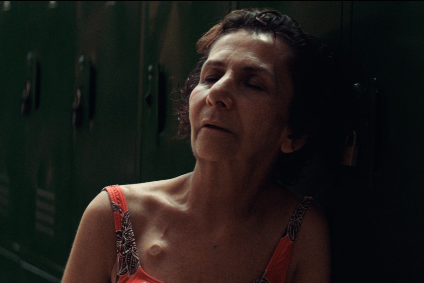 An older woman with dark hair sits in bright and floral swimming costume leaning back on dark green lockers with eyes closed.