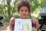 A little girl with curly brown hair smiles at the camera showing her painting on paper