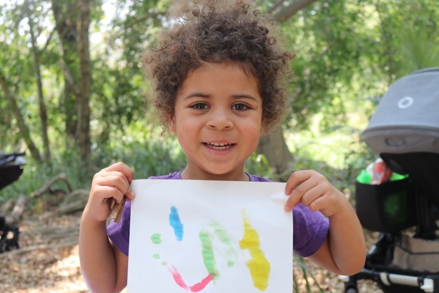 A little girl with curly brown hair smiles at the camera showing her painting on paper