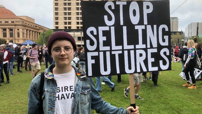 A 14 year old girl in a denim jacket and maroon-coloured beanie holds a handmade sign which says: "stop selling futures".