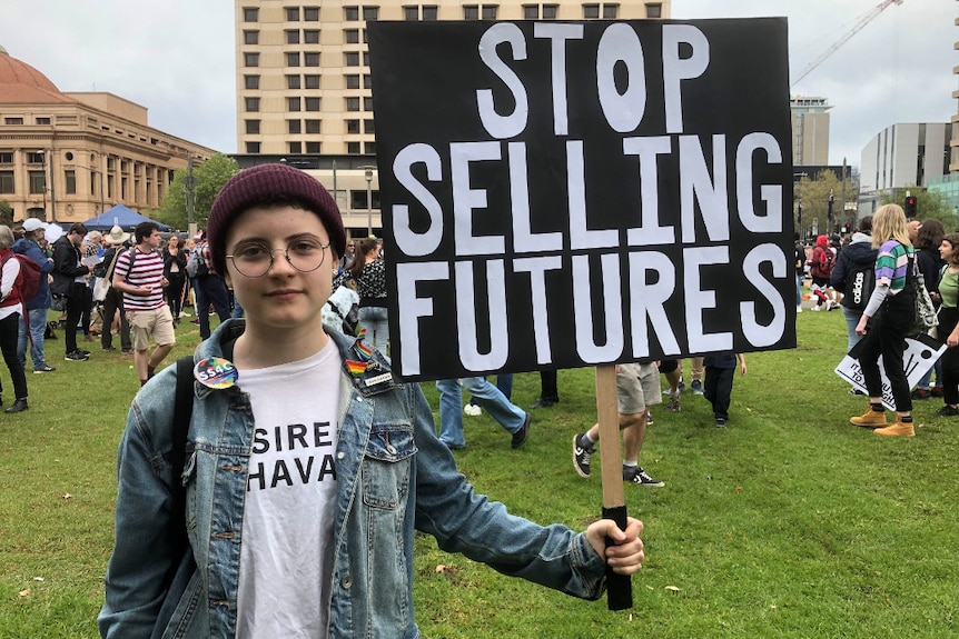 A 14 year old girl in a denim jacket and maroon-coloured beanie holds a handmade sign which says: "stop selling futures".