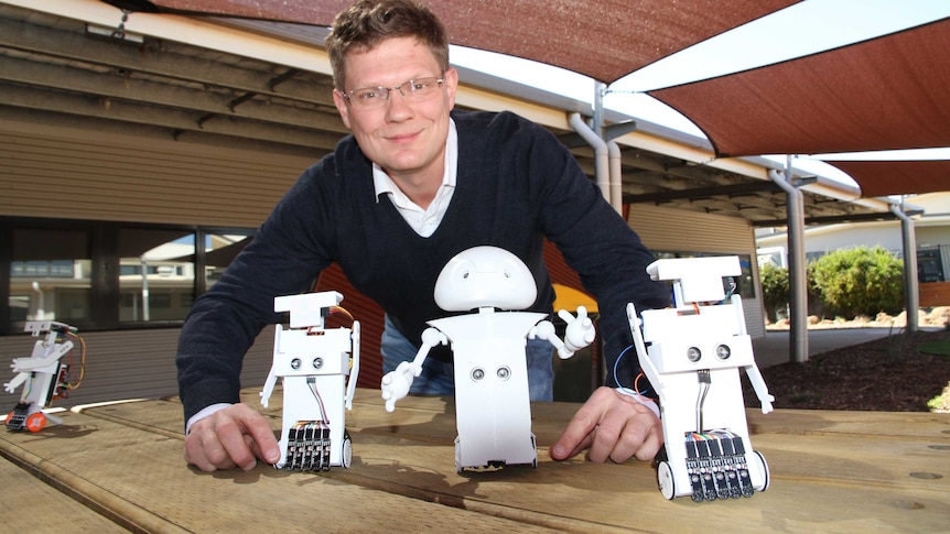 Dr Simon Egerton smiles at camera with three small, white robots in front of him. They're in a school yard.