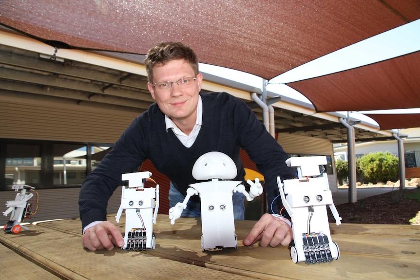 Dr Simon Egerton smiles at camera with three small, white robots in front of him. They're in a school yard.