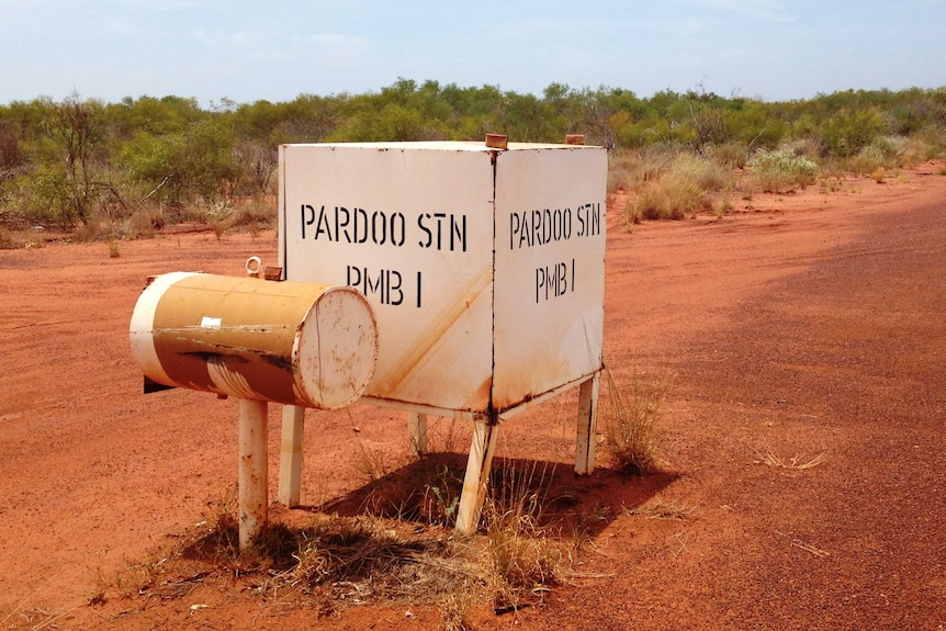 A white mailbox planted in red dirt, with shrubs behind.