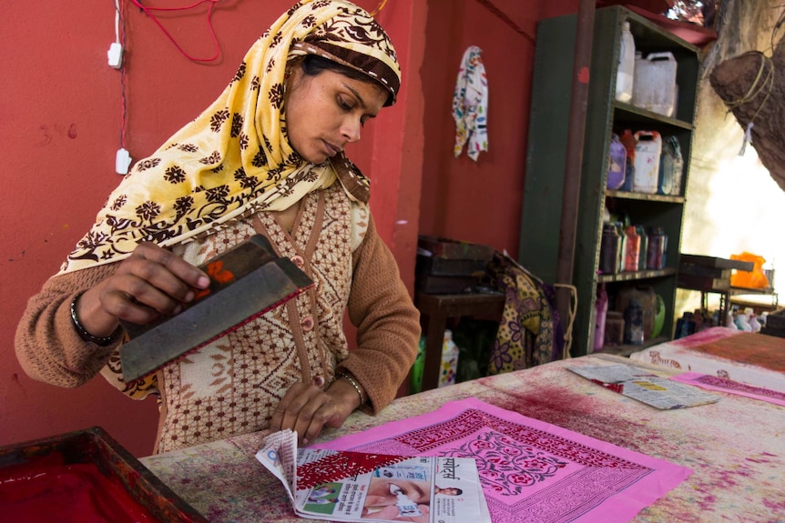 A woman in a yellow headscarf stamps patterns on pink fabric.