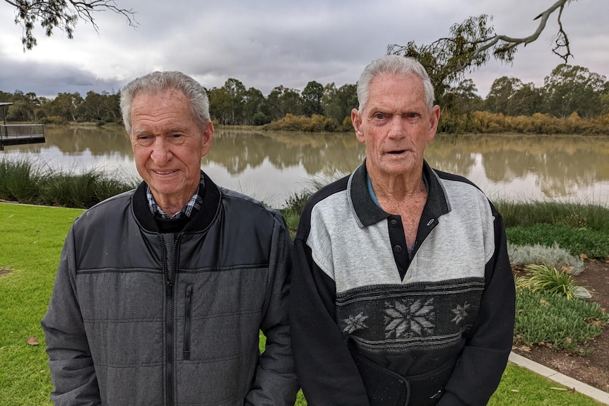 Two men, wearing black jumpers, stand in front of a river. 