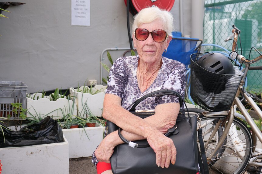 A white-haired woman with sunglasses, a patterned shirt and red bike shorts sits with a black handbag on her lap.