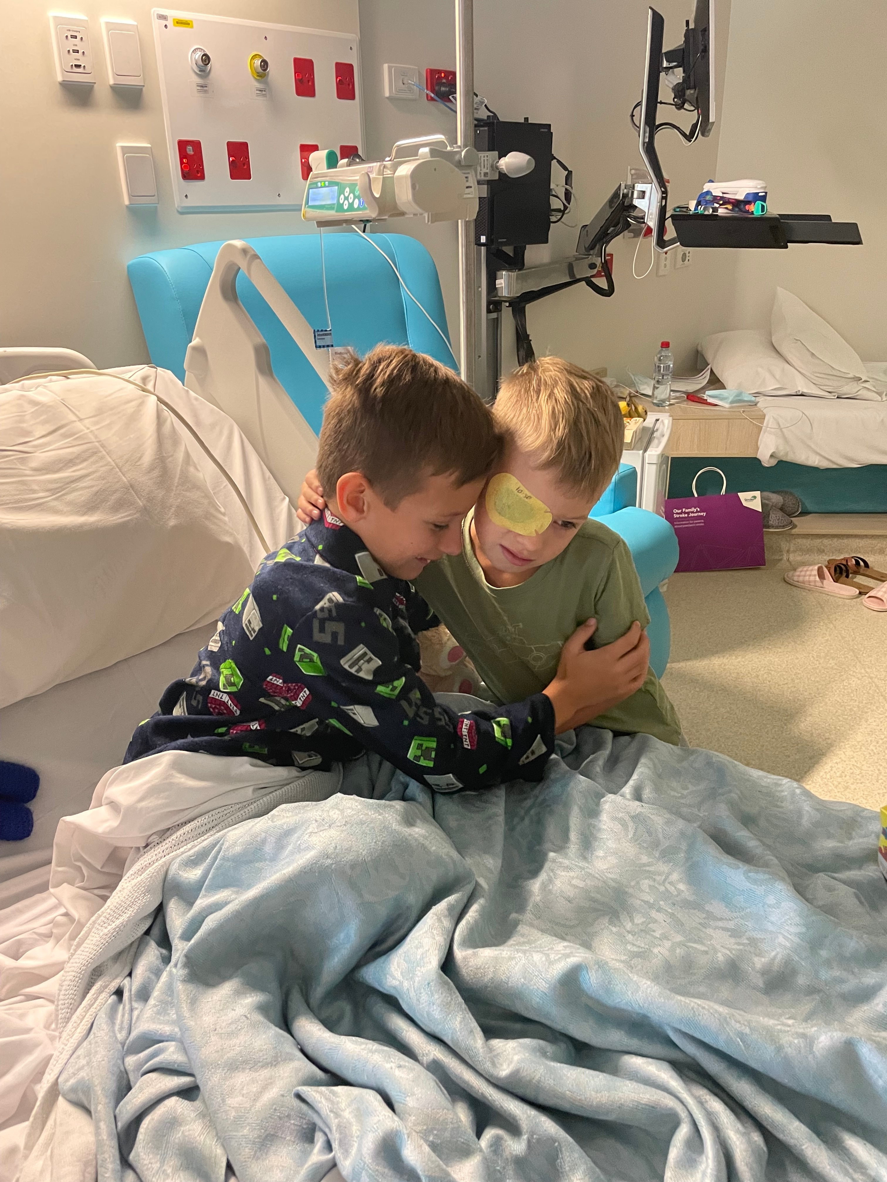 Dos niños pequeños se abrazan en una habitación de hospital. Uno está acostado en una cama de hospital mientras el otro está de pie y lleva un parche en el ojo.