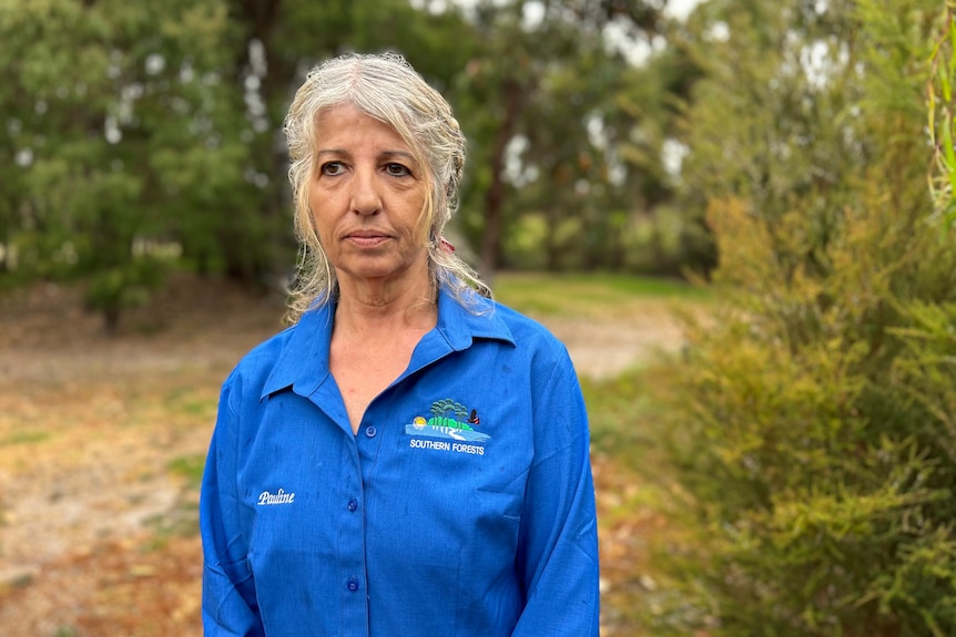 A mid shot of a woman with a blue shirt and grey hair standing outside in front of trees, looking serious.