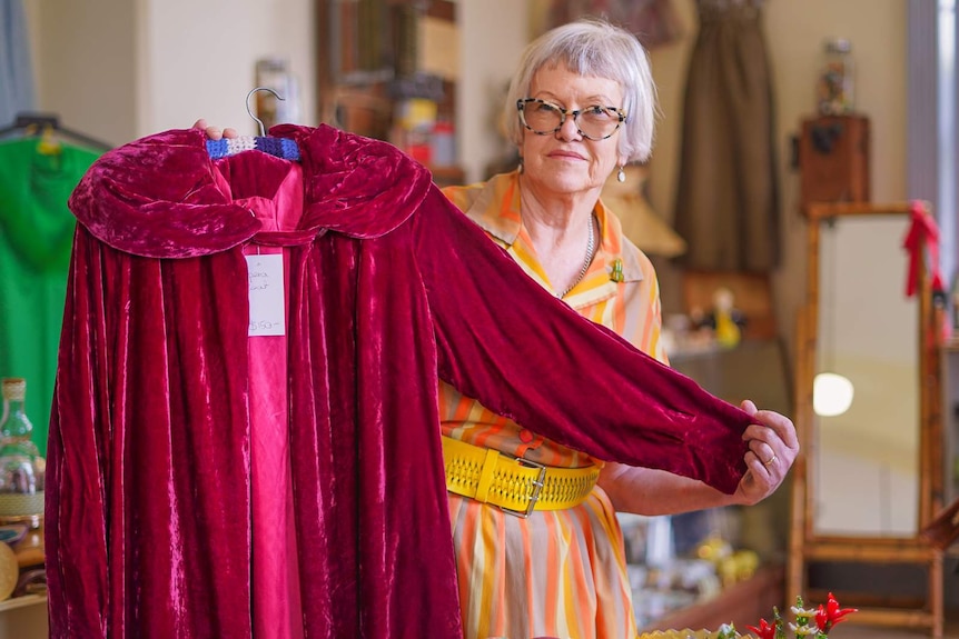 A woman with a grey bob haircut stands holding a red opera cape on a hanger.