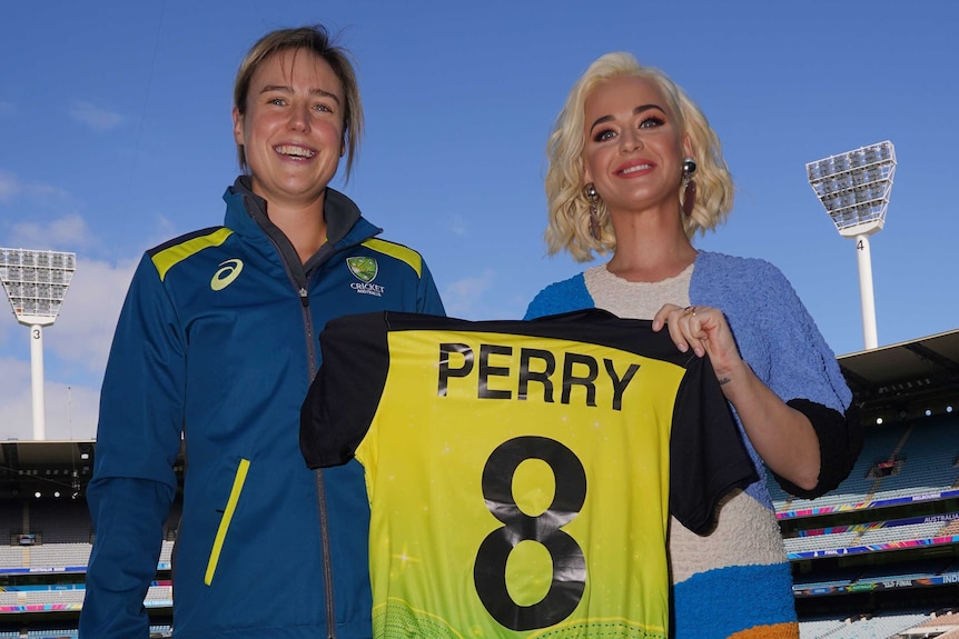 Ellyse Perry and Katy Perry smile while holding up an Australian jersey with 'PERRY 8' on the back.
