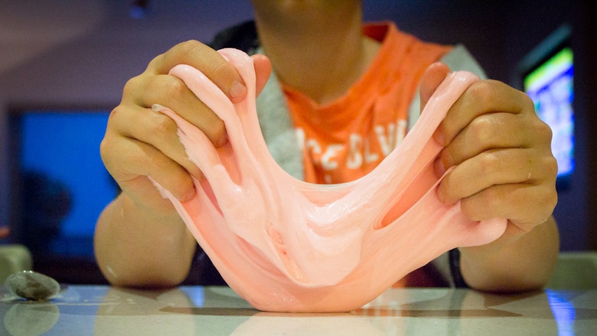 Pink slime being held between a child's hands at a kitchen bench.