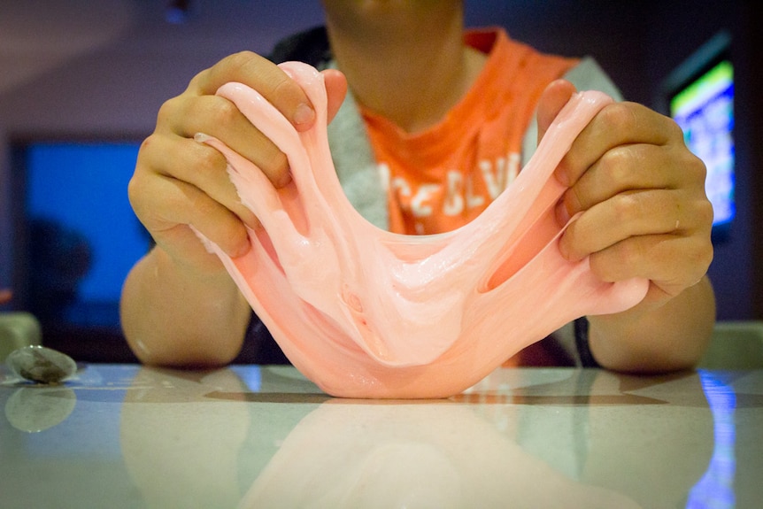 Pink slime being held between a child's hands at a kitchen bench.