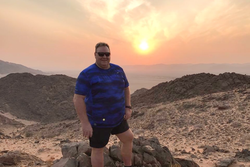 A man wearing a blue and black shirt and shorts stands on a mountain as the sun sets behind him.