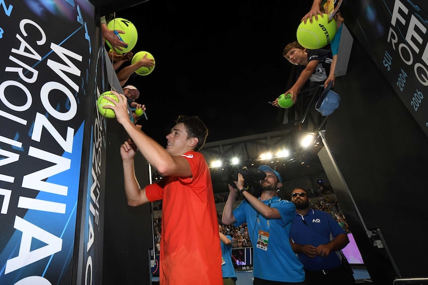Alex de Minaur signs giant novelty tennis balls that fans hang over the stands as a cameraman tracks his movements.