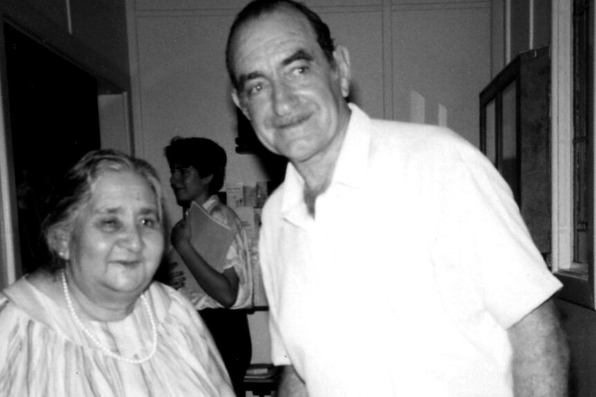 A black and white photo smiling man wearing a white shirt leans close to a short, plump smiling woman wearing strand of pearls.