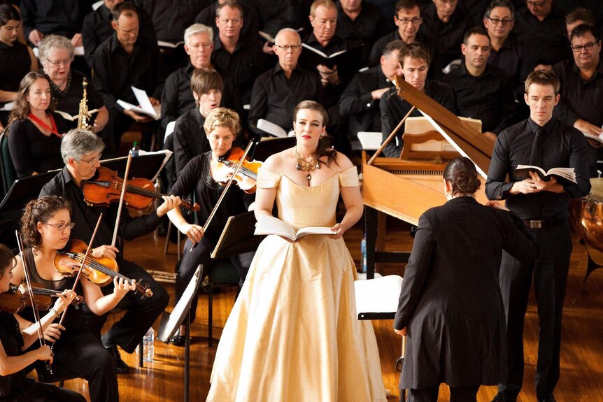 An opera singer wears a gown. She is surrounded by an orchestra wearing black.