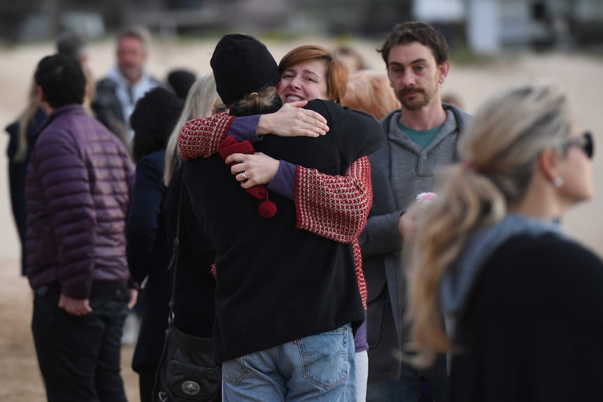 Two people hug each other on a beach at dawn, with a crowd in the background.