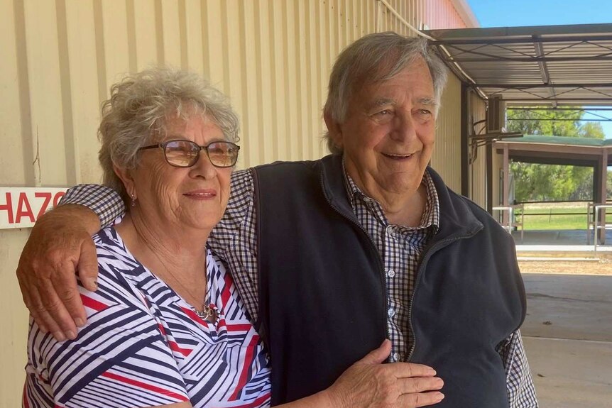 A woman with grey hair and a man with grey hair, smiling outside a steel clad building on a hot and sunny day.