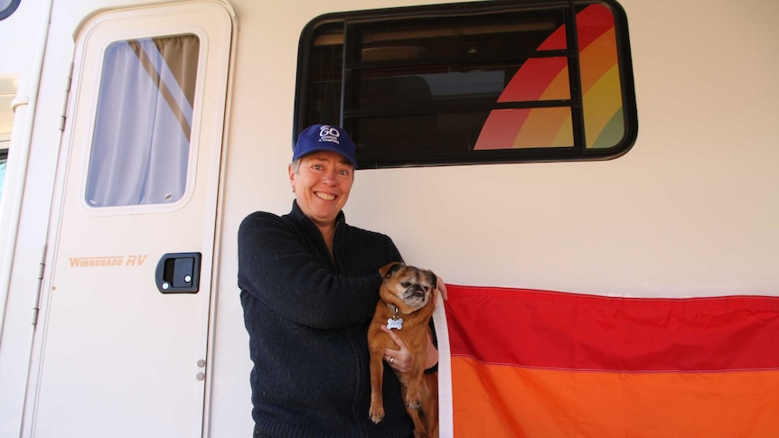 Lynne stands out the front of her caravan with a dog in one arm and an LGBT flag in another