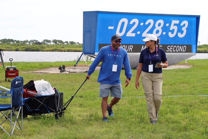 Członkowie mediów odchodzą z trawiastego obszaru w pobliżu miejsca startu rakiet NASA, w tle zatrzymuje się duży zegar odliczający.