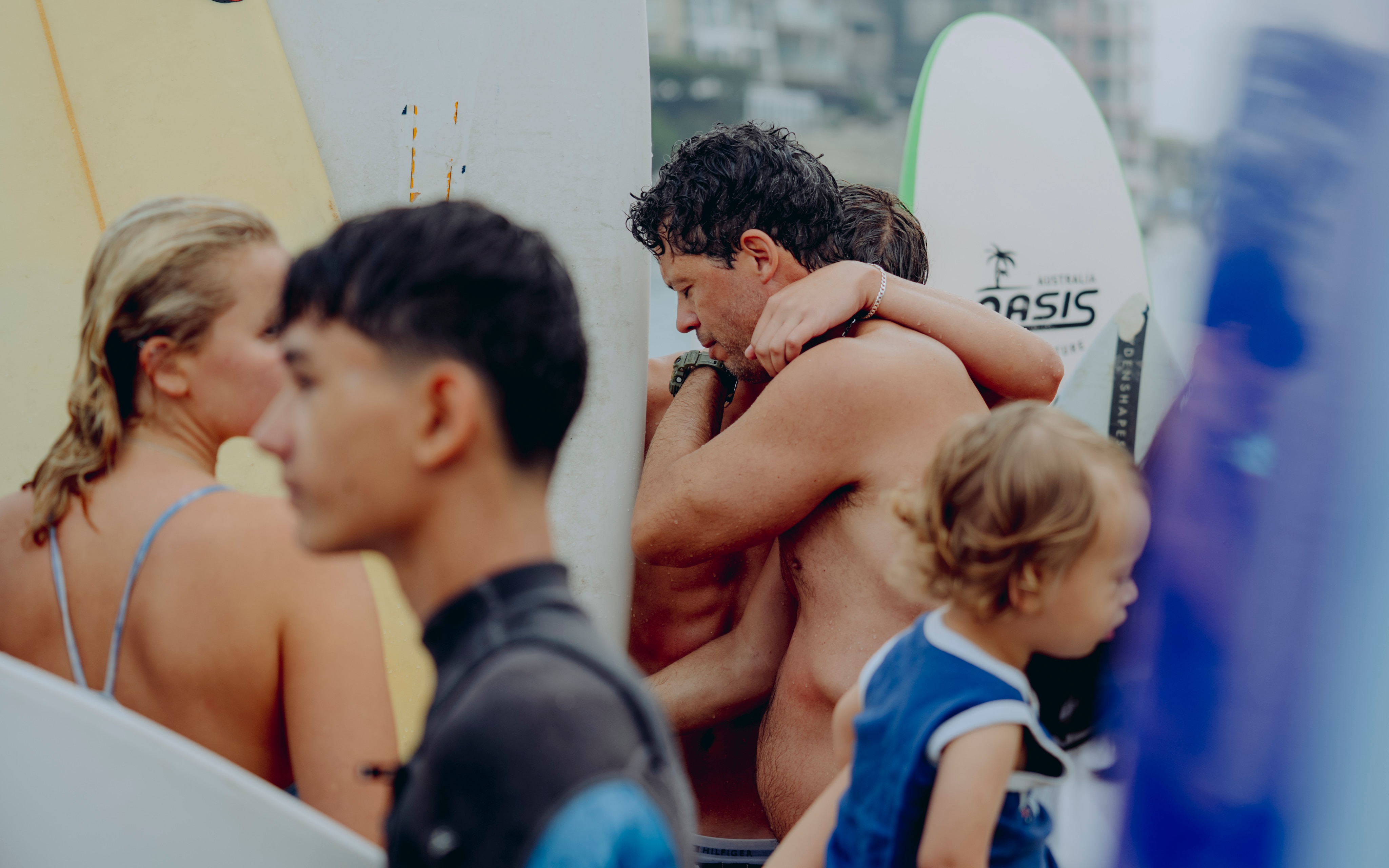 Dois homens são vistos abraçados no meio de uma multidão de pessoas com pranchas de surf e paddle na praia.