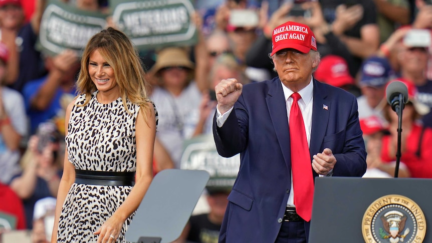 A woman in a leopard pattern dress stands next to a man wearing a suit with a red tie wearing a Make America Great Again cap
