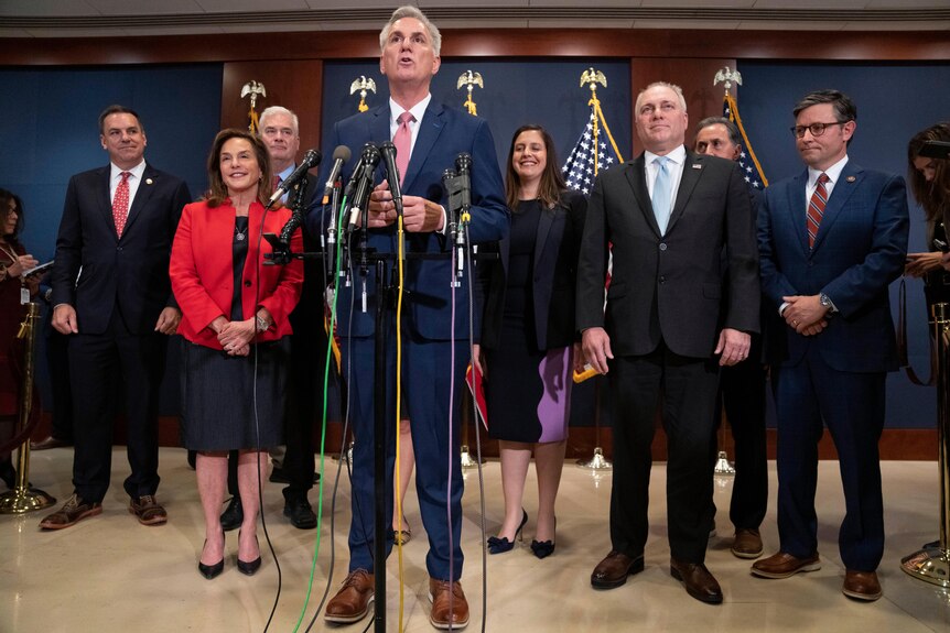Un uomo bianco con un abito blu si trova di fronte a un gruppo di persone sorridenti in giacca e cravatta durante una conferenza stampa.