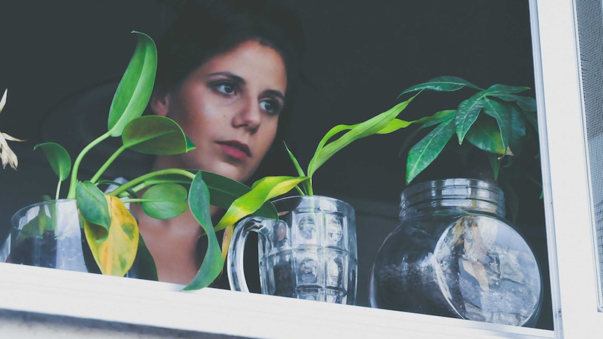 A woman stands at a window with plants growing from jars.