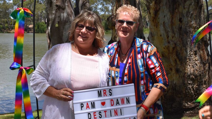 Dawn and Pam Destini holding a sign introducing their married name standing in front of rainbow ribbons and a river.