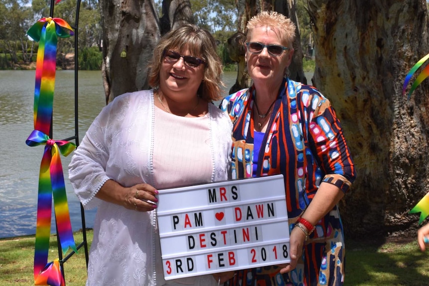 Dawn and Pam Destini holding a sign introducing their married name standing in front of rainbow ribbons and a river.