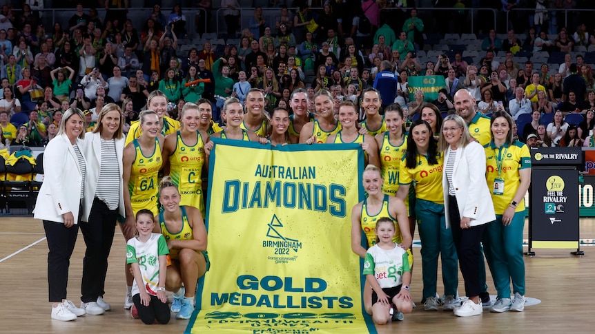 The Australian netball team, staff and coaches smile as they pose with a banner saying 'Australian Diamonds - gold medallists".