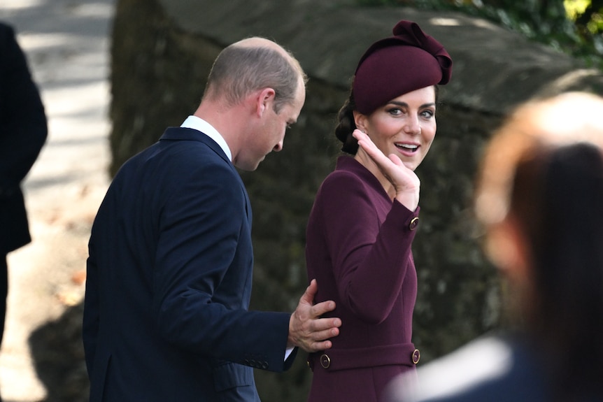 A woman in a burgandy dress and hat waves as a man puts a hand on her back 