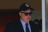 A man in a cap and sunglasses leaves out of a building's glass door.