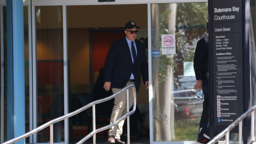 A man in a cap and sunglasses leaves out of a building's glass door.