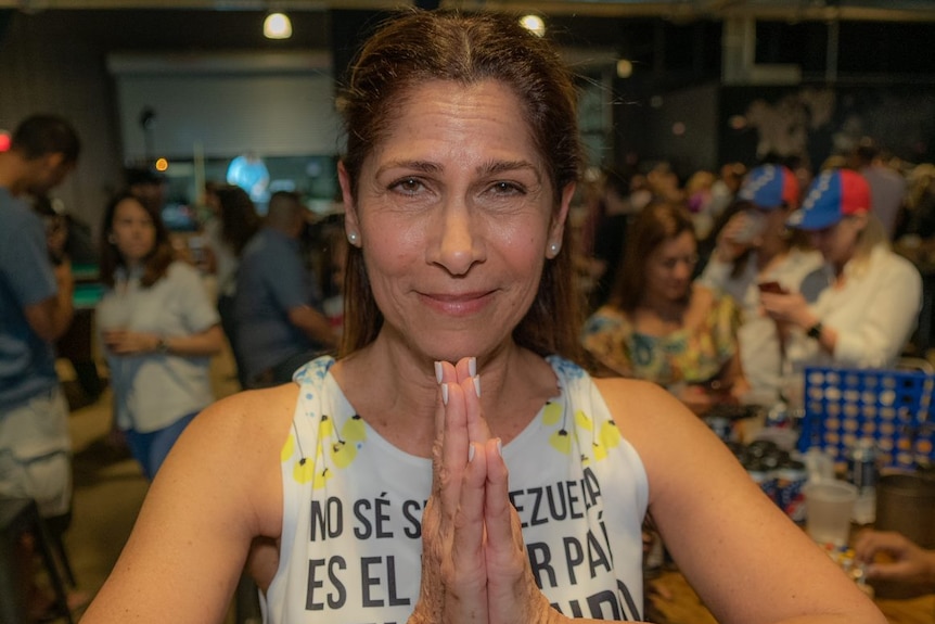 A woman stands with her hands in prayer position at an event, smiling into the camera, with a group of people behind her