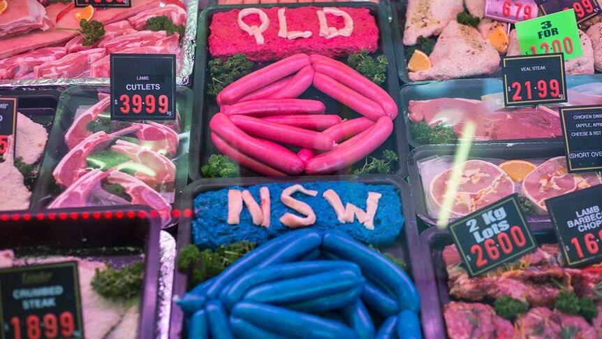 Man with beaming smile, holds two trays of sausages out, one maroon, one blue, with QLD, NSW spelt out at the top in pork rind