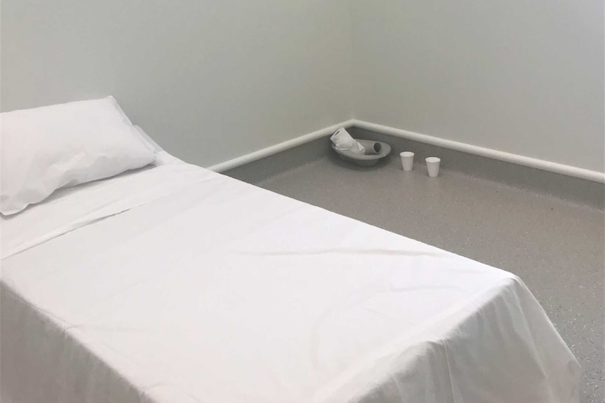 A white mattress on the ground in a grey room, with a CCTV camera on the ceiling and a roll of toilet paper in the corner.