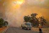 Smoke fills the sky and landscape as two firefighters stand near a fire truck  on a road and another sprays bushland.
