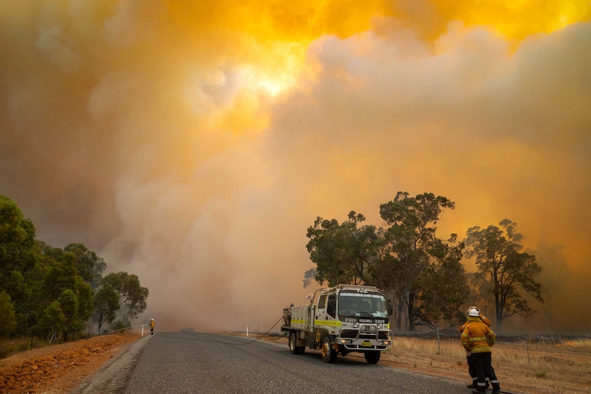Smoke fills the sky and landscape as two firefighters stand near a fire truck  on a road and another sprays bushland.