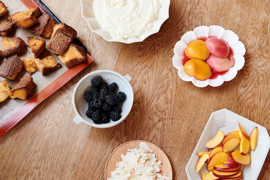 Trifle ingredients on a timber table: fresh peach slices, coconut pieces, blackberries and pandoro pieces.