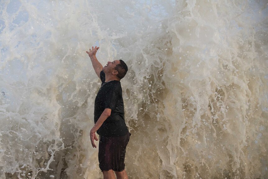 A man stands under a wave as it hits a seawall.