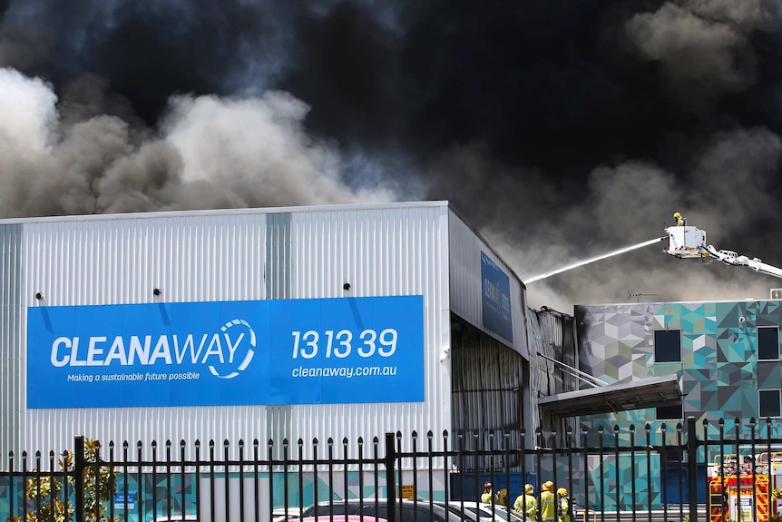 Black smoke fills the sky above a Cleanaway warehouse as a firefighter in a fire truck basket sprays water on the building.