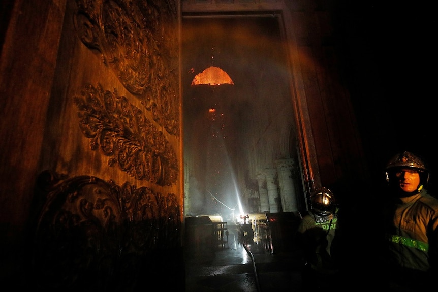 Firefighters enter a burning cathedral through an ornate entrance and set up equipment to battle the blaze