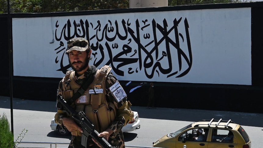 A Taliban fighter in a military uniform stands in front of a black and white mural with Arabic calligraphy