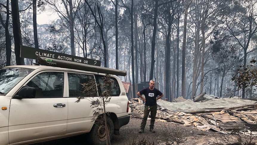 A man stands next to a car that has a home made sign on the roof saying "Climate action now...?"  He is surrounded by burnt bush