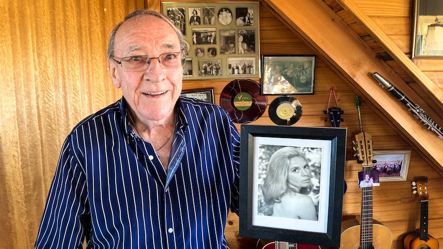 A smiling man in his 80s holds a black and white photo of a beautiful young woman.