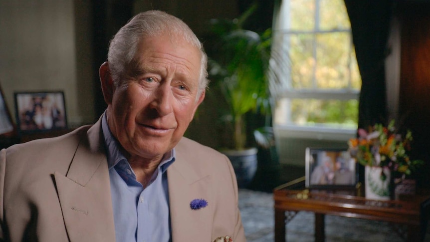 Prince Charles sits in a living room, with family photos in the background.